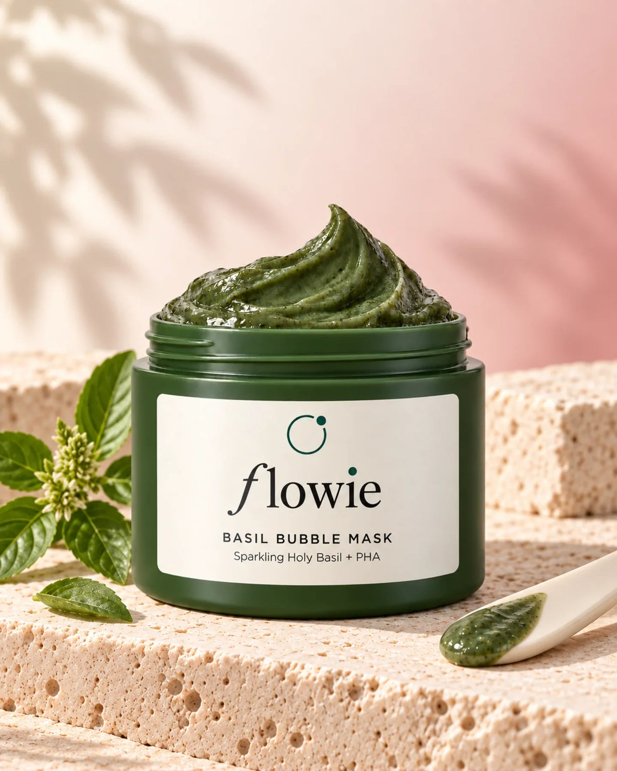 Open Basil Bubble Mask jar with basil leaves and a cream ceramic spatula on pale stone